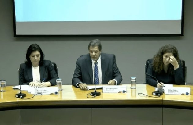 Cinco pessoas sentadas em mesa de conferência. Um homem fala ao microfone. Visíveis placas de nome e bandeira do Brasil.