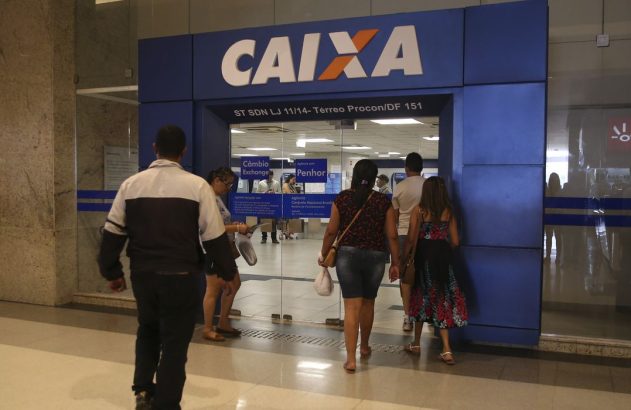 Pessoas em frente à agência da CAIXA, com letreiro do banco e serviços como Câmbio e Penhor.
