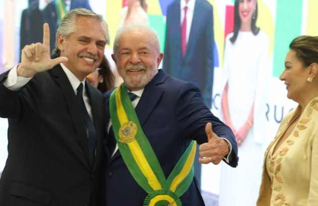 Alberto Fernández e Lula sorrindo. Fernández faz sinal de 'L', Lula, de faixa presidencial, polegar. Mulher observa.