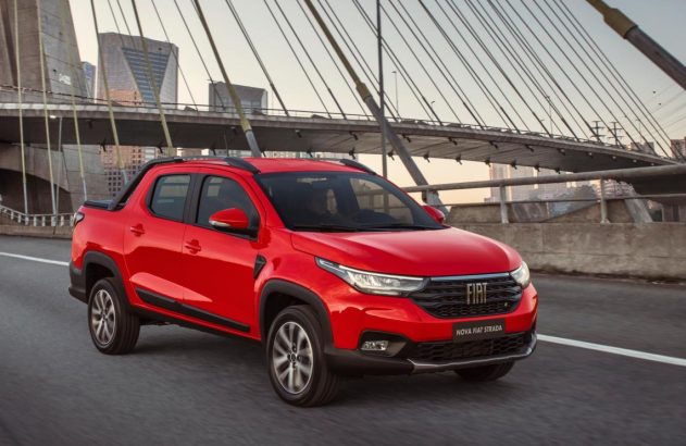 Picape Fiat Nova Strada vermelha rodando em estrada urbana, com ponte e prédios ao fundo.