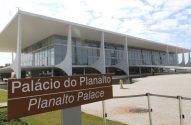 Vista frontal do Palácio do Planalto, edifício modernista branco com fachada de vidro, sob céu azul. Placa indica "Palácio do Planalto".