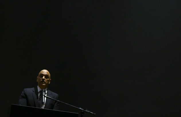 Homem careca de terno escuro discursa num púlpito iluminado, em fundo preto.