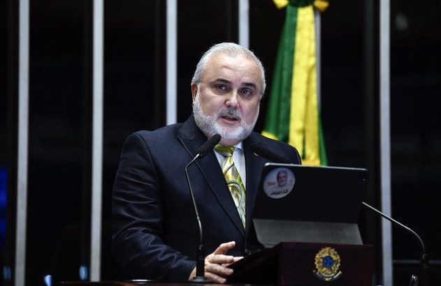 Homem de terno e barba branca discursa em púlpito oficial, com bandeira brasileira ao fundo.