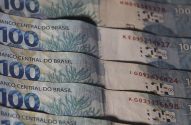 Pilha de notas de 100 Reais brasileiros, com "Banco Central do Brasil" e o valor "100" visíveis.