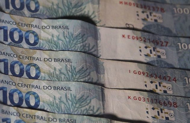 Pilha de notas de 100 Reais brasileiros, com "Banco Central do Brasil" e o valor "100" visíveis.