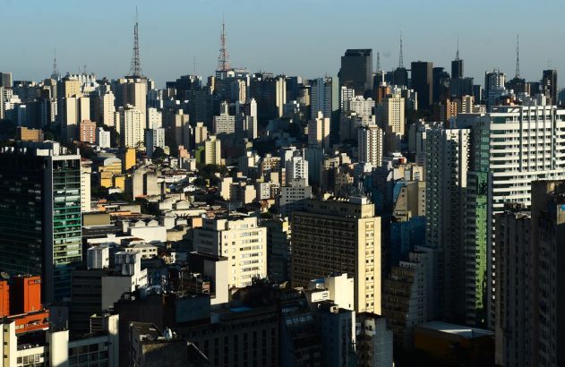 Paisagem urbana densa com inúmeros edifícios altos. Torres de comunicação se erguem acima do horizonte de prédios sob céu azul.