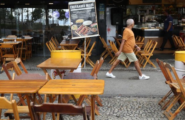Homem caminha em calçada com mesas e cadeiras de madeira de restaurante. Uma placa de churrasco exibe preços.