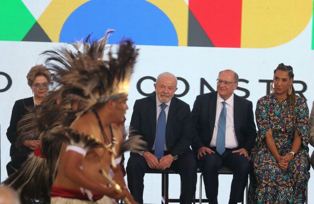 Lula, Alckmin e Dilma observam performance indígena. Tela com texto 'CONSTRUI' ao fundo.