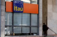 Fachada de agência bancária Itaú com logo azul e amarelo. Uma mulher caminha para a entrada do prédio.