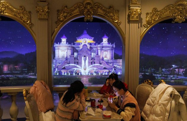 Três mulheres sentadas à mesa em salão elegante, olhando celulares. Ao fundo, uma projeção noturna de castelo e céu estrelado.