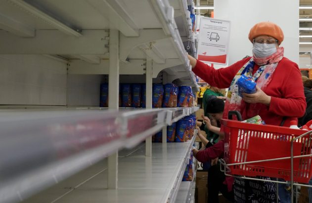 Mulher mascarada compra em supermercado com prateleiras esvaziadas. Sinal russo "Mercadoria a caminho".