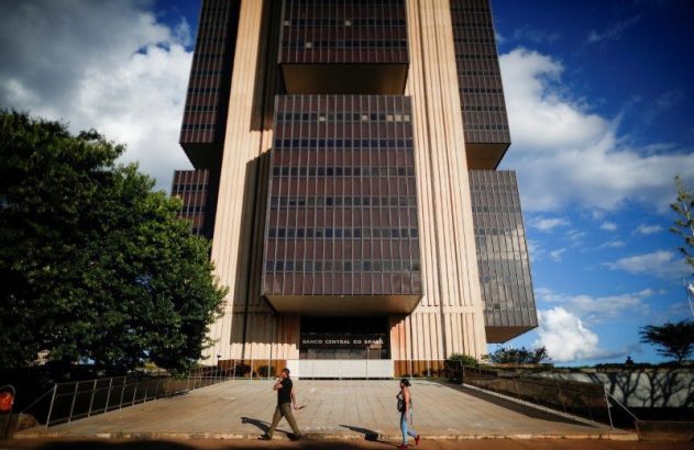 Fachada do Banco Central do Brasil sob céu azul, com duas pessoas caminhando na rampa de acesso.