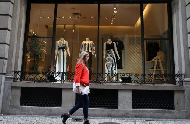Mulher de jaqueta vermelha anda em frente à vitrine de loja com manequins e roupas listradas.