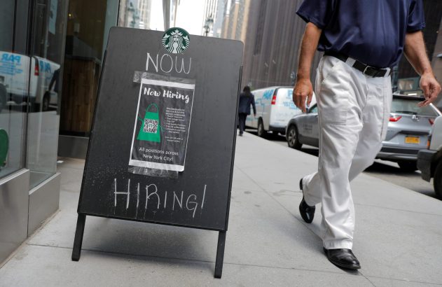 Placa de 'Contratando Agora' do Starbucks, com QR code, e um homem caminhando em calçada urbana.