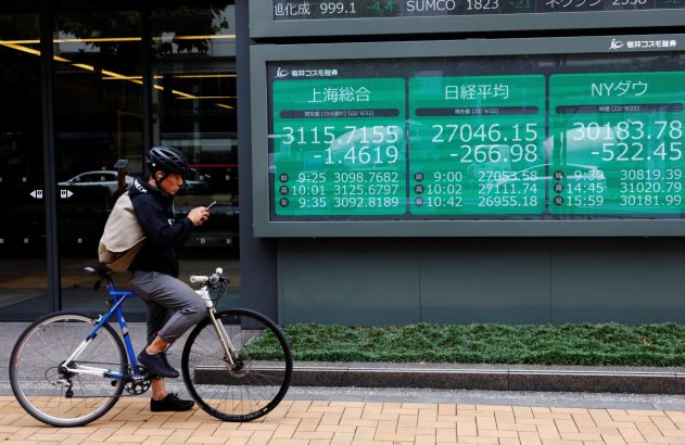 Um ciclista olha o smartphone ao lado de um painel digital exibindo cotações da bolsa de valores.