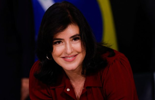 Mulher de cabelos escuros e camisa vinho sorri. Fundo desfocado azul e amarelo.