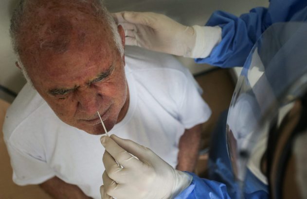 Profissional com luvas e avental azul faz teste nasal em homem idoso. Ele está com os olhos fechados enquanto cotonete é inserido.