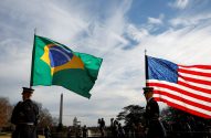 Duas bandeiras (Brasil e EUA) hasteadas, com soldados de guarda e o Obelisco de Washington ao fundo. Céu azul com nuvens.