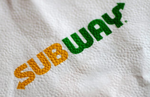 Logo da Subway com setas, em verde e amarelo, sobre guardanapo branco texturizado.