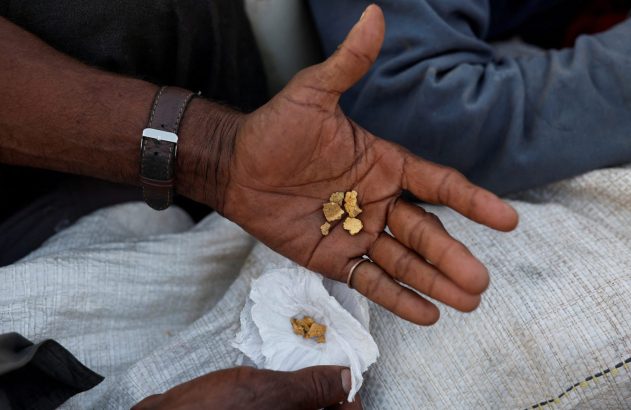 Mão aberta exibe pepitas de ouro na palma. Outra mão segura ouro em papel sobre um saco branco.