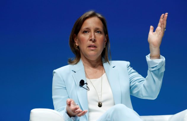 Mulher de terno azul claro sentada, discursando e gesticulando. Fundo azul liso.