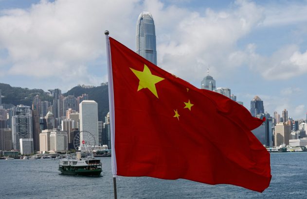 Bandeira da China em primeiro plano, com o horizonte de Hong Kong, incluindo arranha-céus e um ferry, ao fundo.