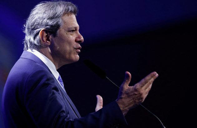 Homem de terno azul e cabelo grisalho fala em microfone, gesticulando.