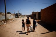Mulher e duas crianças caminham em rua de terra, carregando galão de água e garrafa.