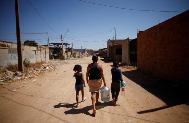 Mulher e duas crianças caminham em rua de terra, carregando galão de água e garrafa.