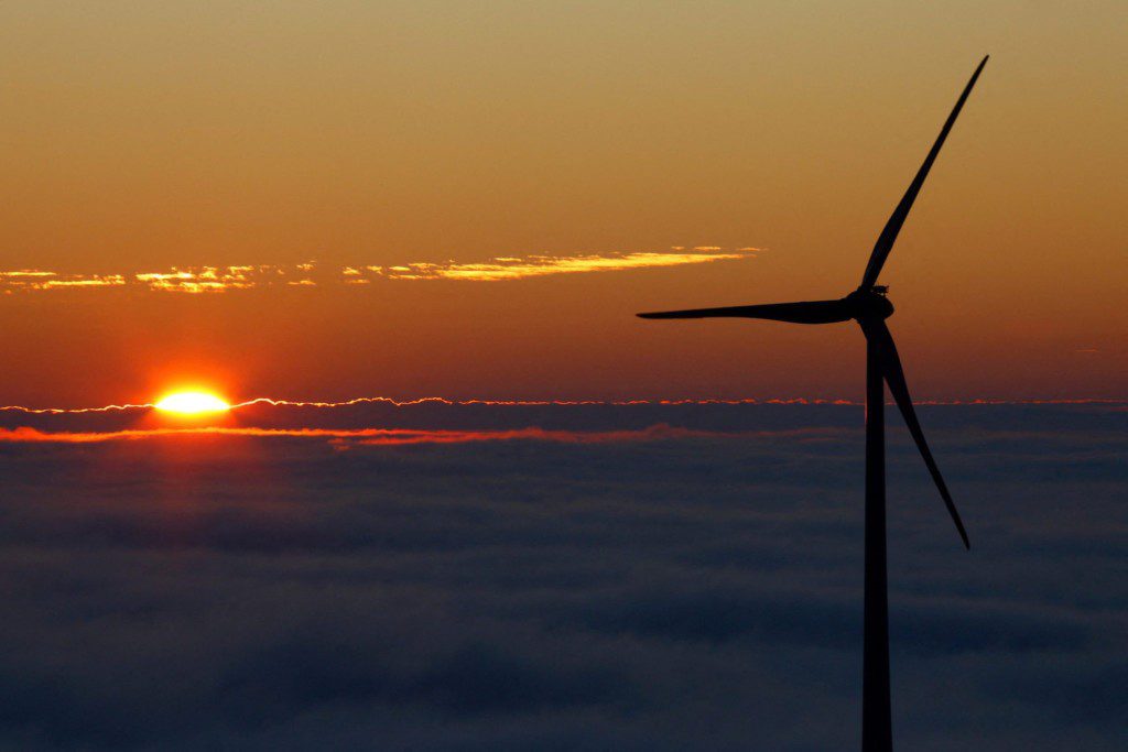 Turbina eólica em silhueta e sol se pondo sobre um mar de nuvens.