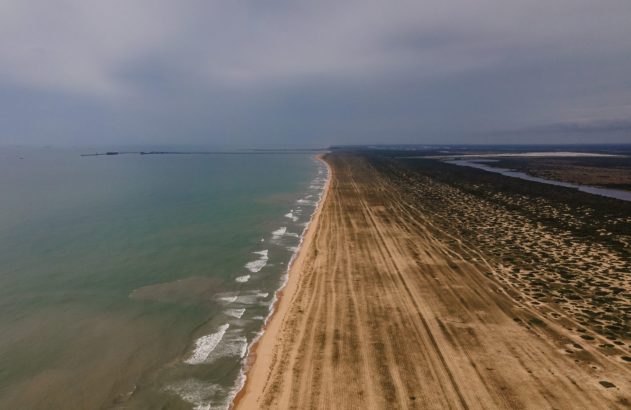 Longa faixa de terra entre o oceano (com praia e ondas) e um rio, com trilhos e vegetação, sob céu nublado.