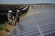 Três trabalhadores inspecionam painéis solares em campo. Um usa medidor. Vasta usina de energia limpa sob céu azul.