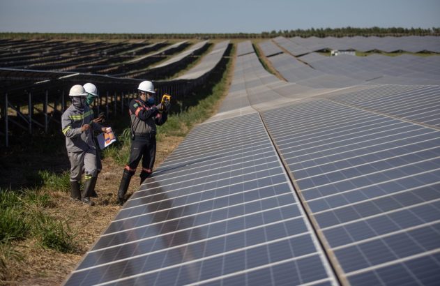 Três trabalhadores inspecionam painéis solares em campo. Um usa medidor. Vasta usina de energia limpa sob céu azul.