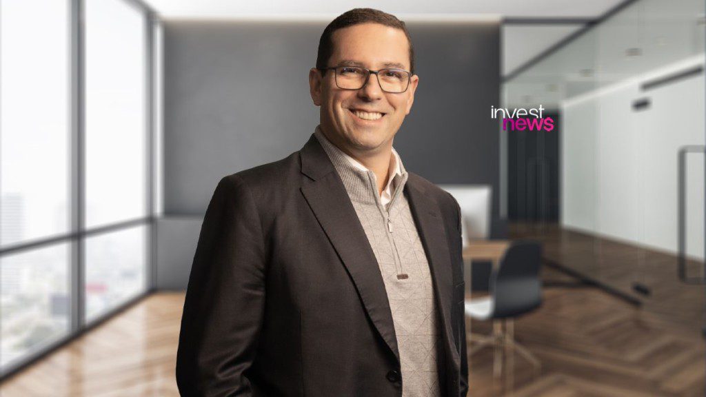 Homem de óculos sorri. Veste blazer e suéter quadriculado em escritório moderno. Fundo com logo "invest news".