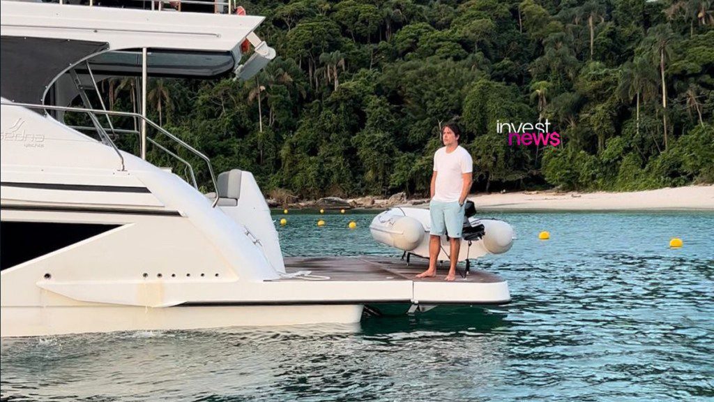 Homem em pé na plataforma de um iate branco, com mata e praia ao fundo.