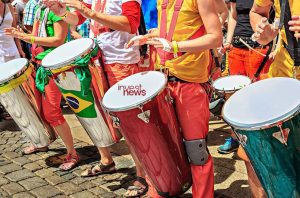 Pessoas tocam tambores. Alguns com bandeira do Brasil, um com logo 'invest news'.