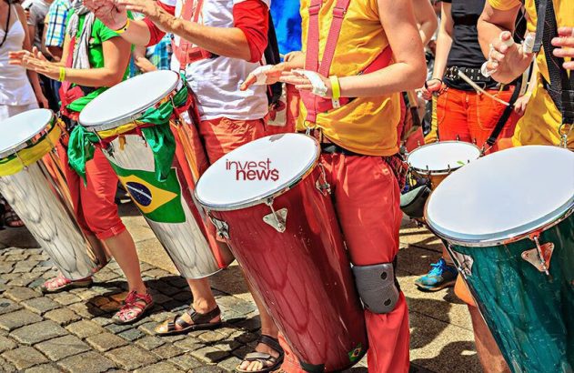 Pessoas tocam tambores. Alguns com bandeira do Brasil, um com logo 'invest news'.