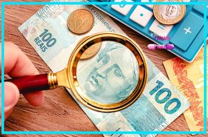 Lupa examinando uma nota de 100 reais, com calculadora e moedas ao fundo.
