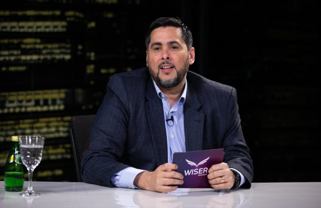 Homem em terno, com microfone, fala em estúdio. Segura folheto Wiser Educação sobre mesa com água. Fundo escuro com luzes.