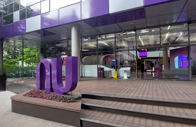 Logo roxo 'nu' do Nubank na entrada de um prédio moderno. Fachada roxa e cinza, com deck de madeira