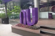 Um grande letreiro roxo "NU" na entrada de um edifício moderno com fachada de vidro e jardim.
