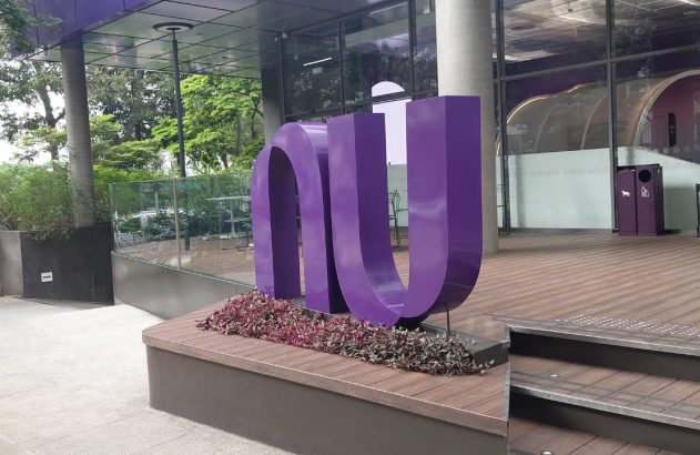 Um grande letreiro roxo "NU" na entrada de um edifício moderno com fachada de vidro e jardim.