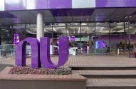 Grande logo roxo "NU" em frente a prédio de vidro com deck de madeira.