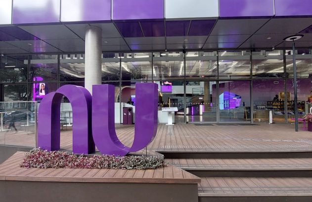 Grande logo roxo "NU" em frente a prédio de vidro com deck de madeira.