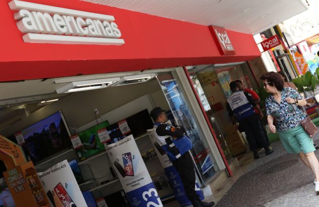Fachada da loja Americanas e local, com pessoas na calçada, um segurança e eletrônicos à mostra.