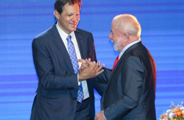 Fernando Haddad segura a mão de Lula com as duas mãos, ambos sorrindo.