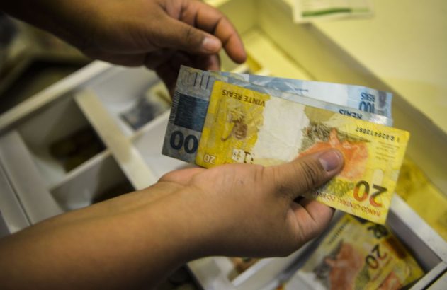 Mãos seguram e separam cédulas de 20 e 100 reais, sobre um organizador de dinheiro branco.