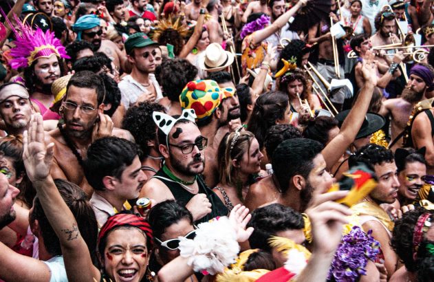 Uma multidão diversa e animada celebra um festival. Pessoas fantasiadas, alguns tocam instrumentos, outros levantam as mãos.
