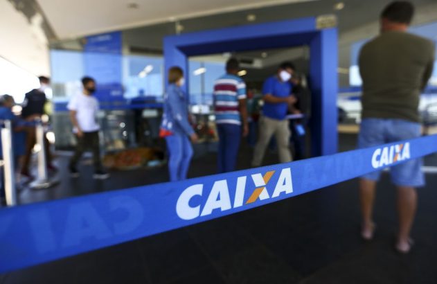 Fila de pessoas em frente a uma agência bancária, com uma fita azul da "CAIXA" em primeiro plano.