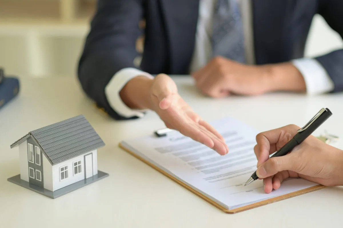 Mão de uma mulher segurando uma caneta e assinando um contrato de financiamento. À sua frente um homem de terno aponta para o documento. Ao lado uma miniatura de uma casa.